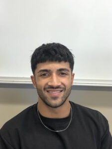 A man with short dark hair and a beard is wearing a black shirt and a chain necklace, smiling against a plain white background.