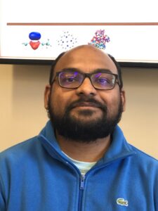 Person wearing glasses and a blue fleece jacket poses in front of a screen displaying molecular diagrams.