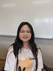 A person with long hair and glasses sitting in a classroom with a whiteboard in the background, wearing a T-shirt with a graphic and carrying a backpack.