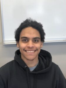 Person with curly hair smiling in front of a whiteboard, wearing a dark hoodie.