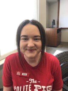 A person wearing a red shirt and name tag, seated indoors. A window and a desk are visible in the background.