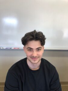 A person with dark hair and a black shirt sits in front of a whiteboard, smiling slightly.
