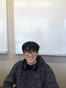 A person wearing glasses and a hoodie sits in front of whiteboards, smiling with headphones around their neck.