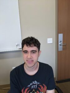 A person with short dark hair is sitting in a room with a whiteboard and a closed door. They are wearing a black T-shirt with a red and yellow graphic design.