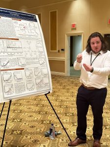 A person in a white shirt presents a scientific poster on a stand in a conference room. The room has beige walls, patterned carpet, and an exit sign above a door in the background.
