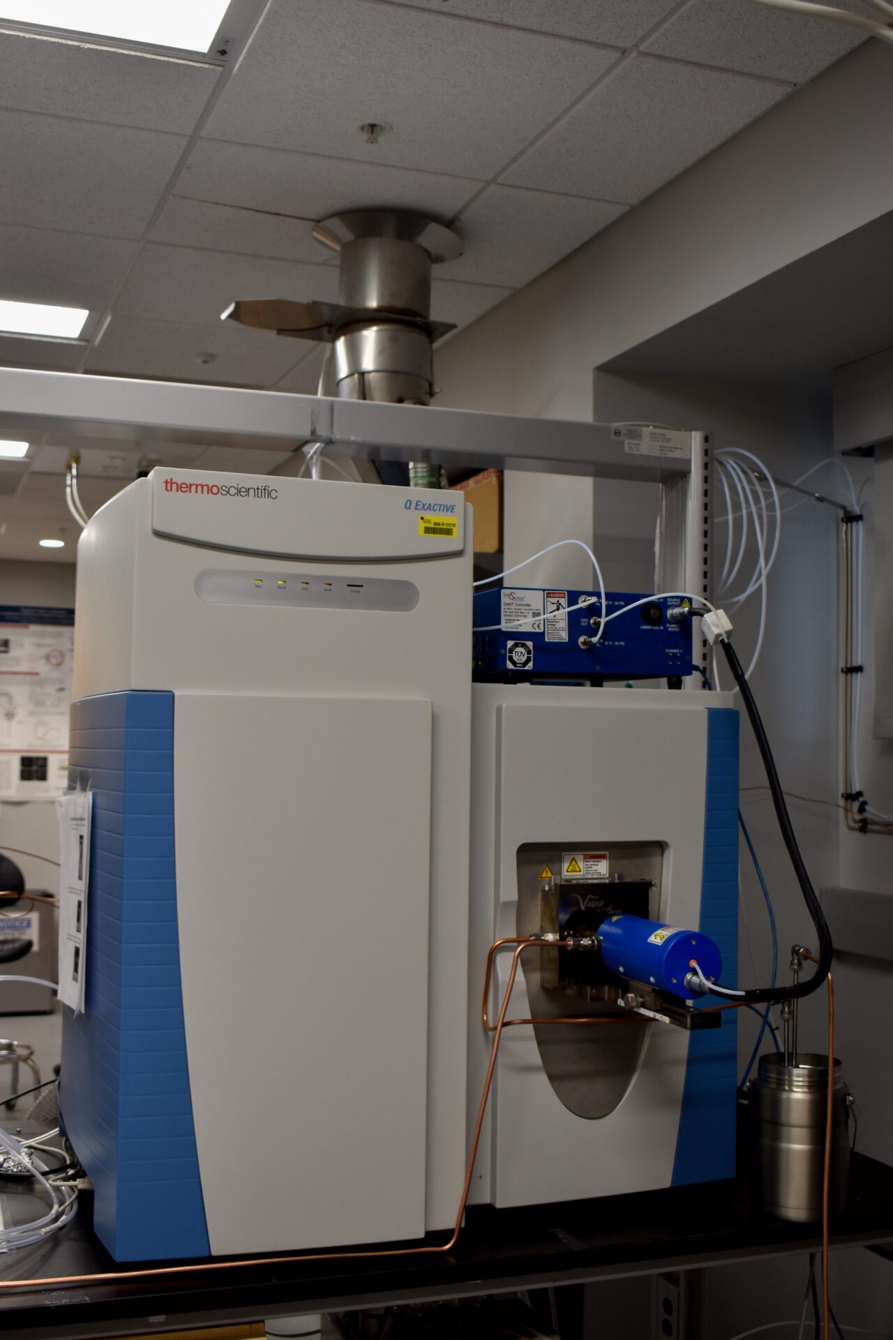 A large scientific instrument with blue and gray casing, featuring cables and a small display, situated in a laboratory setting under fluorescent lights.