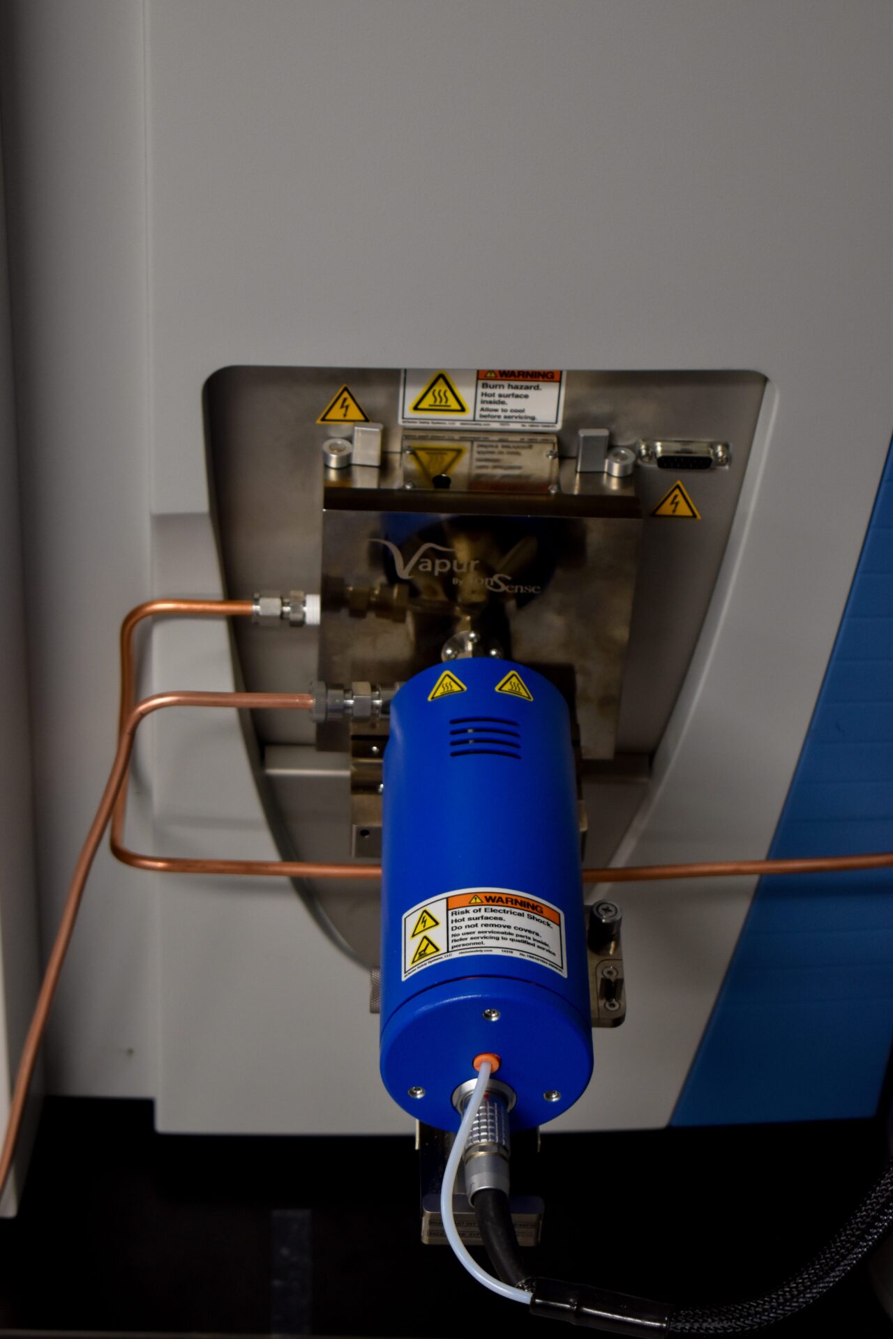A laboratory instrument with a blue component and copper tubes, featuring several caution labels.