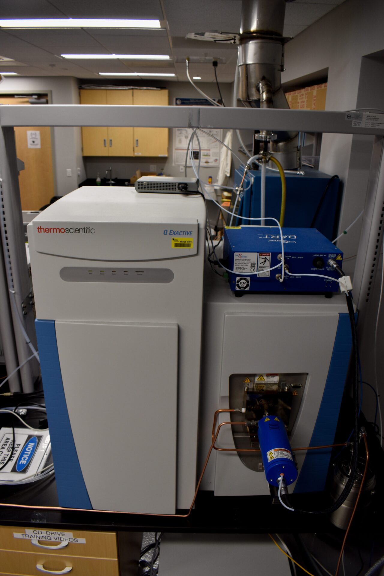 A laboratory mass spectrometer setup with various connected components, including a "Thermo Scientific" unit and a blue analyzer. Shelving and lab cabinets visible in the background.