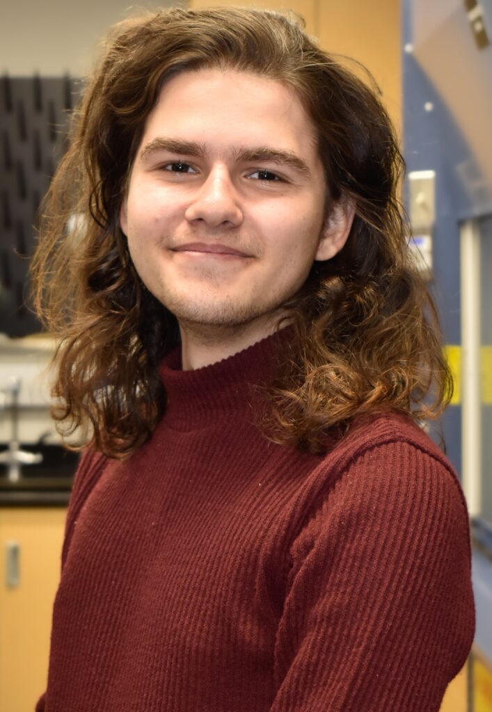 A person with long brown hair is smiling while wearing a maroon sweater. They're in a room with blurred background elements.
