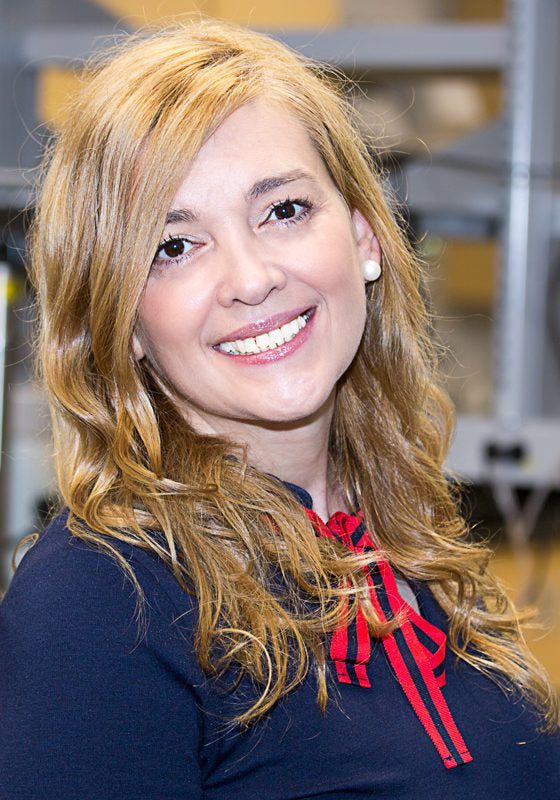 Smiling person with long, wavy blonde hair wearing a navy top with red accents, in an indoor setting.
