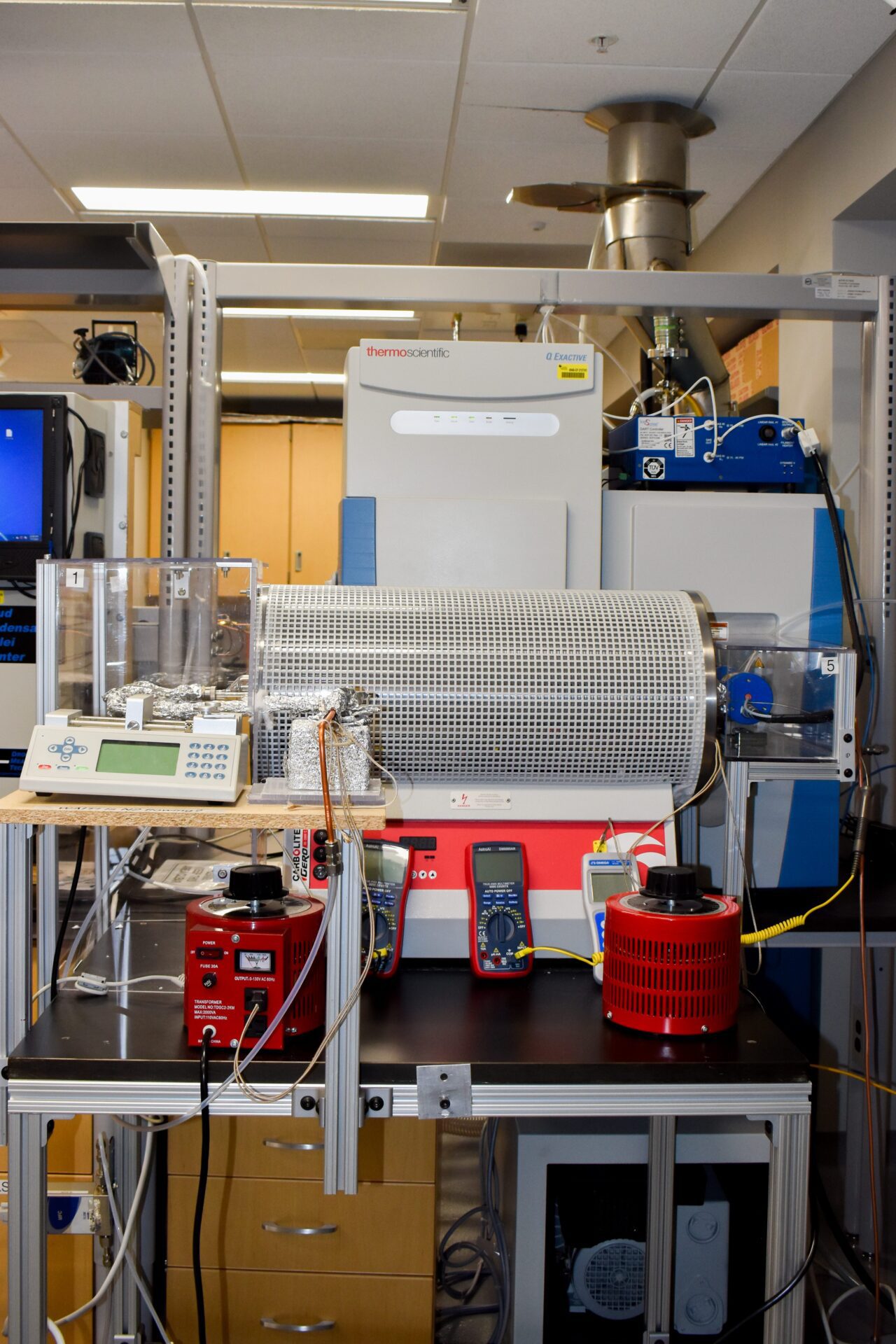 A laboratory setup featuring a horizontal tube furnace, digital multimeters, a scale, and various electronic equipment on a workbench.