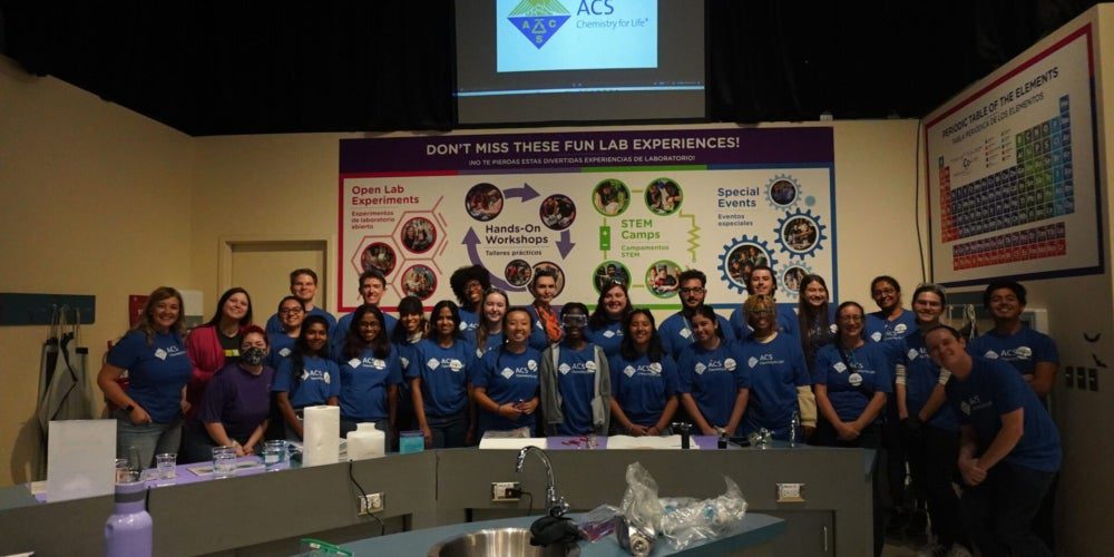 A group of people in matching blue ACS t-shirts poses for a group photo in a science lab with educational posters and equipment visible.