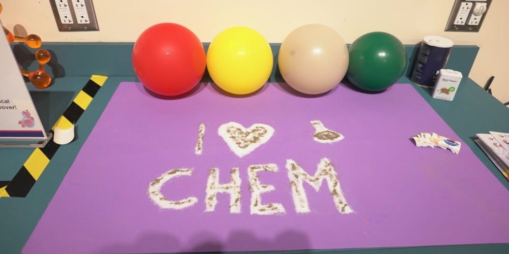 A purple mat with white powder spelling "I ♥ CHEM" and drawing of a flask, four balloons, and science materials on a countertop.