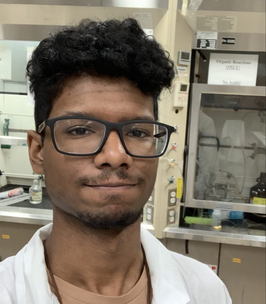 A person in a laboratory wearing a white lab coat and glasses stands in front of lab equipment, looking at the camera.