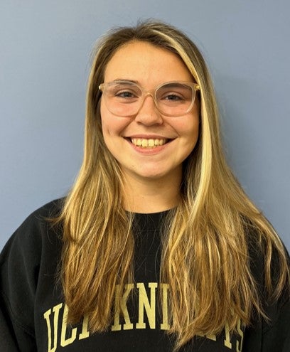 A young woman with long blonde hair, wearing glasses and a black "UCF Knights" sweatshirt, smiles in front of a plain light blue background.