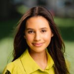 A young woman with long brown hair wearing a yellow-green collared shirt smiles at the camera outdoors, with a blurred green background.