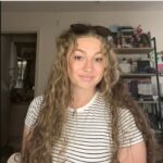 A woman with long curly hair wearing a white and black striped shirt sits indoors in front of shelves and a doorway, smiling slightly at the camera.