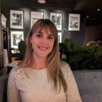 A woman with straight, light brown hair and a light sweater sits in a restaurant with framed black-and-white photos on the wall and green plants behind her.