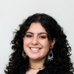 A person with curly dark hair, wearing a black top, dangling earrings, and a necklace, smiles at the camera against a plain light background.