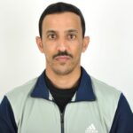A man with short black hair and a mustache wearing a light grey and navy Adidas zip-up jacket, facing the camera against a plain white background.