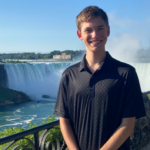 A person stands and smiles in front of a railing with a large waterfall and mist in the background on a sunny day.