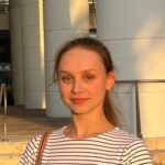 A young woman wearing a white and black striped shirt stands outside in sunlight, with large white columns and steps in the background.