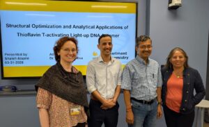Four people stand in front of a presentation slide titled "Structural Optimization and Analytical Applications of Thioflavin T-activating Light-up DNA Aptamer" in a classroom setting.
