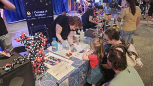 A woman at a UCF College of Sciences booth explains a science activity to a young girl and two adults, with molecular models and educational materials on the table.