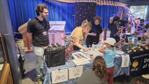 Adults and children interact at a science booth with DNA models, educational materials, and displays, inside a museum-like setting with a large skeleton in the background.