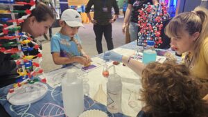 Children and adults interact with science exhibits at a table, featuring DNA models, lab bottles, and hands-on activities at a public event.
