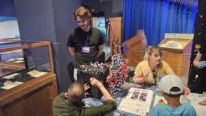 People interact at a science exhibit table featuring a large DNA model, molecular models, and educational materials, with two adults assisting two children.