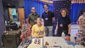 Four people stand and sit behind a table with science models, informational materials, and lab items at a science fair or outreach event. A blue curtain and decorative elements are in the background.