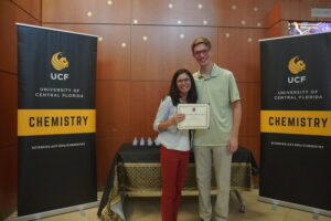 Two people stand smiling between two UCF Chemistry banners, with one person holding a certificate in front of a table with trophies.