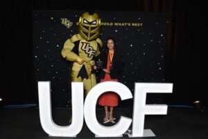 A person holding an award poses next to the UCF knight mascot behind large white UCF letters and a backdrop that reads "BUILD WHAT'S NEXT.