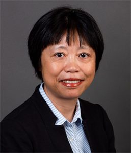 A person with short black hair wearing a dark blazer over a striped shirt, against a plain dark background.