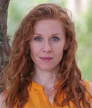 A woman with long curly red hair and blue eyes, wearing a sleeveless yellow top, stands outdoors in front of a tree and blurred greenery.