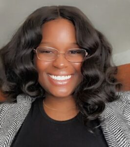 A person with wavy dark hair, wearing glasses and a striped blazer, is smiling at the camera.