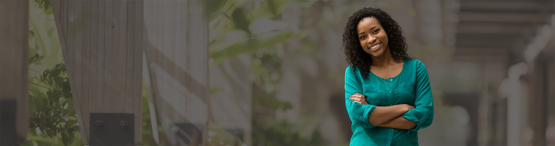 A woman with curly hair, wearing a green top, stands outdoors with arms crossed, smiling at the camera.