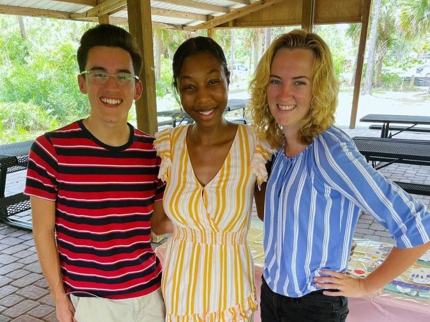 Three smiling friends standing together under a pavilion at UCF, with lush greenery in the background.