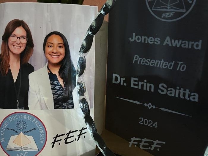 Two women smiling beside an award with the inscription, "Jones Award Presented To Dr. Erin Saitta 2024," representing UCF chemistry education, and a logo for McKnight Doctoral Fellowship and FEF.