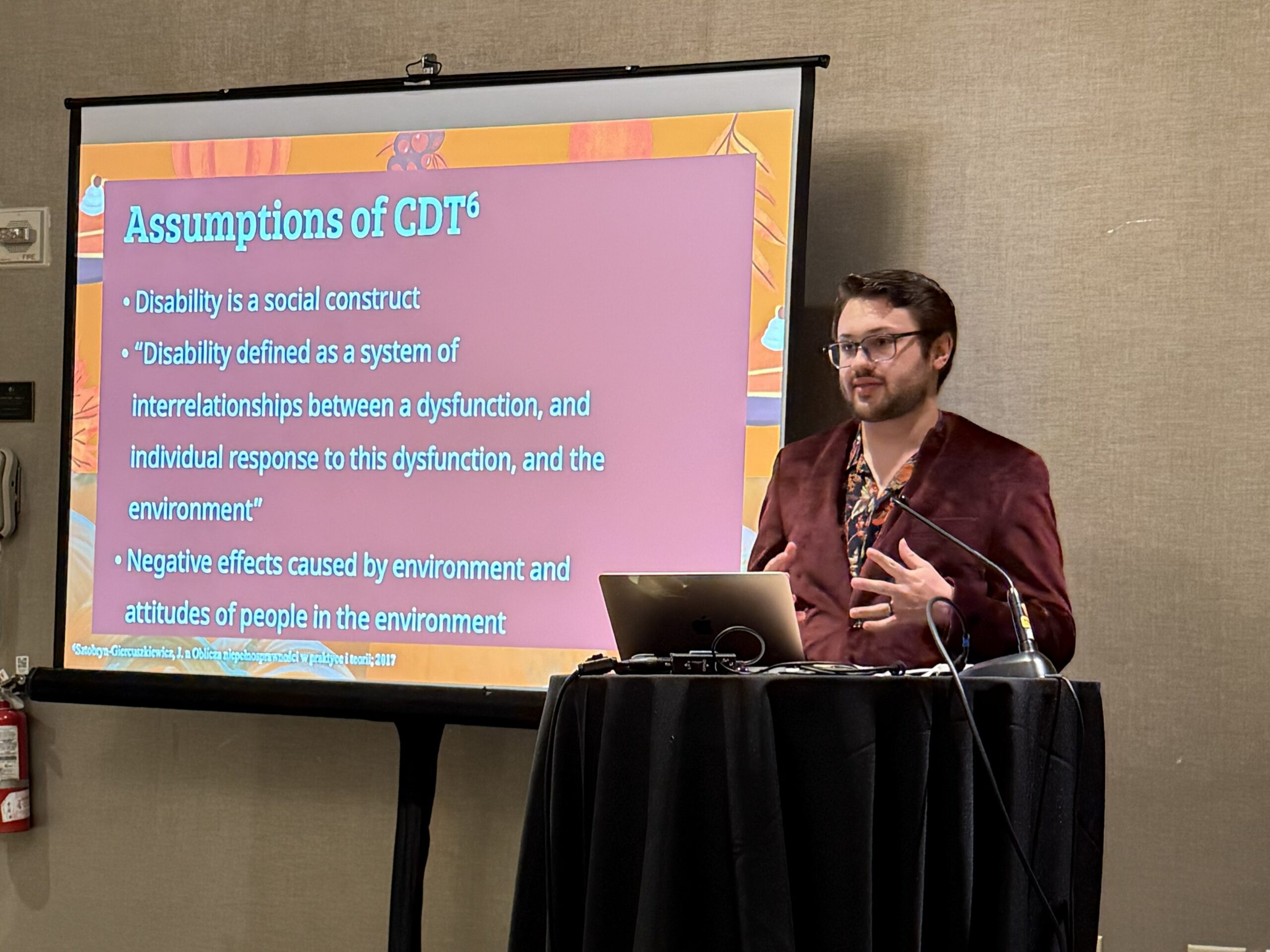 A man presents a slide titled "Assumptions of CDT" discussing disability as a social construct, with a laptop and microphone on a podium—part of a UCF chemistry education seminar.