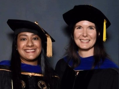 Two women in academic regalia, including black gowns, blue hoods, and doctoral caps with gold tassels, smile proudly against a plain dark background—celebrating their UCF chemistry education achievements.