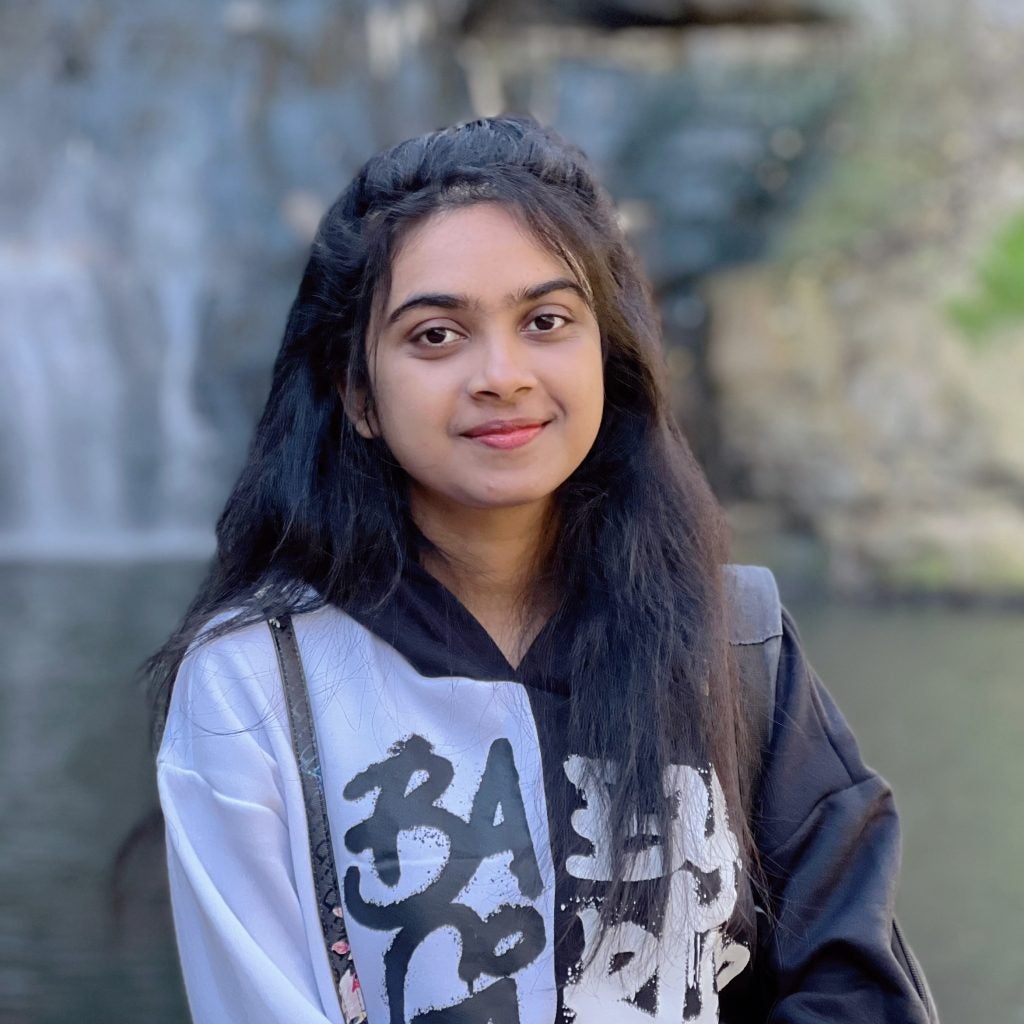 Young woman with long black hair, wearing a black and white hoodie, standing in front of a waterfall and smiling.