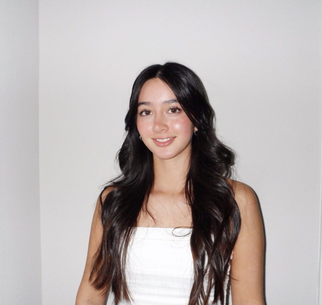 Person with long dark hair, wearing a white strapless top, stands against a plain white background.
