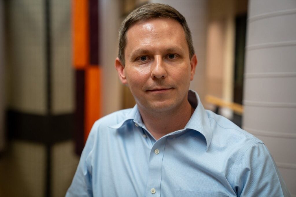 A man in a light blue button-up shirt stands indoors, looking directly at the camera with a neutral expression. The background appears to be an office or hallway.