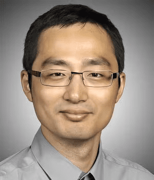 A man with short dark hair, glasses, and a light gray collared shirt, posing in front of a plain gray background.