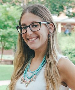 Young woman with long, light brown hair and glasses smiling outdoors, wearing a white sleeveless top and layered turquoise necklaces. Greenery and a building are in the blurred background.