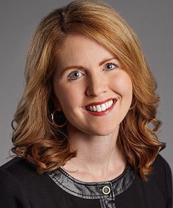 A woman with shoulder-length wavy auburn hair, wearing a black top with leather trim, smiling at the camera against a plain gray background.