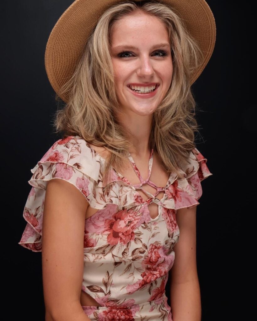 A woman with blonde hair wearing a floral dress and a wide-brimmed hat smiles in front of a black background.