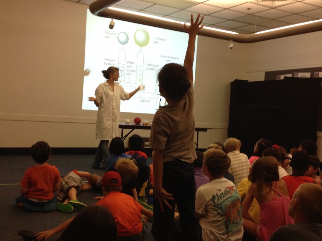 An engaged child raises a hand to ask a question during a science presentation.
